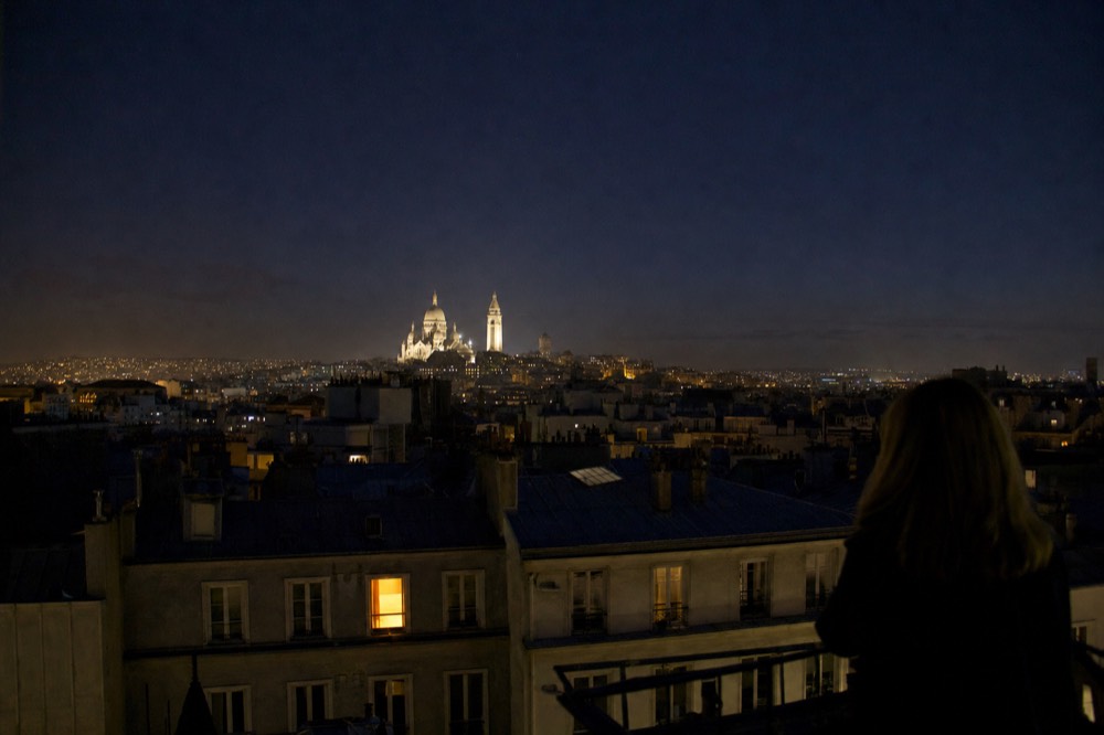 Floodlit Sacre-Coeur rises over dark Paris rooftops at night, with a silhouetted person looking across the skyline.