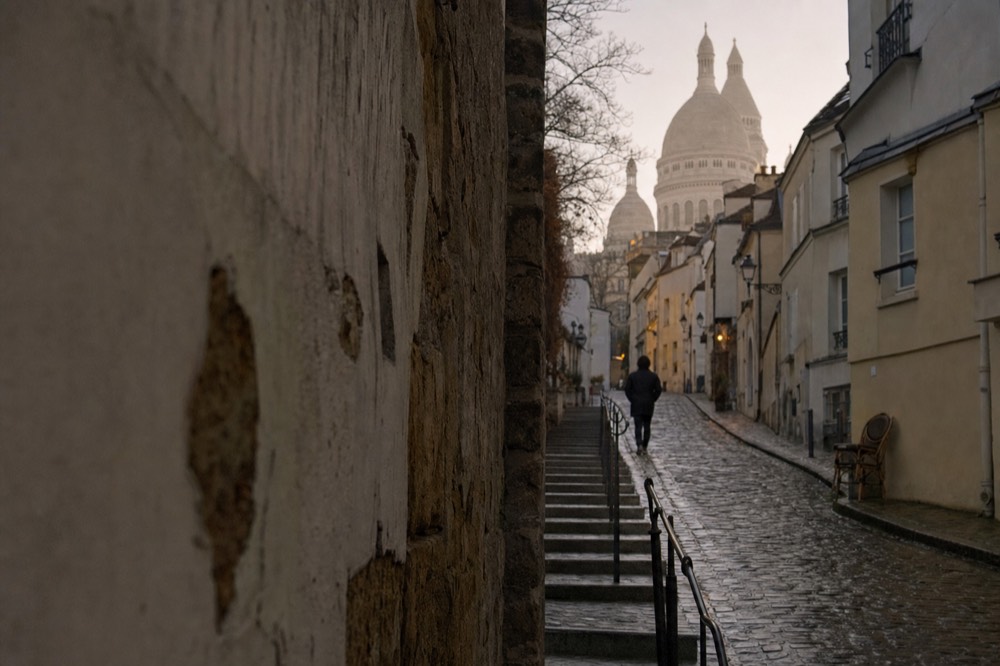 Wet cobbled Montmartre lane and steps lead uphill between pale buildings toward the domes of Sacre-Coeur with a lone distant walker.