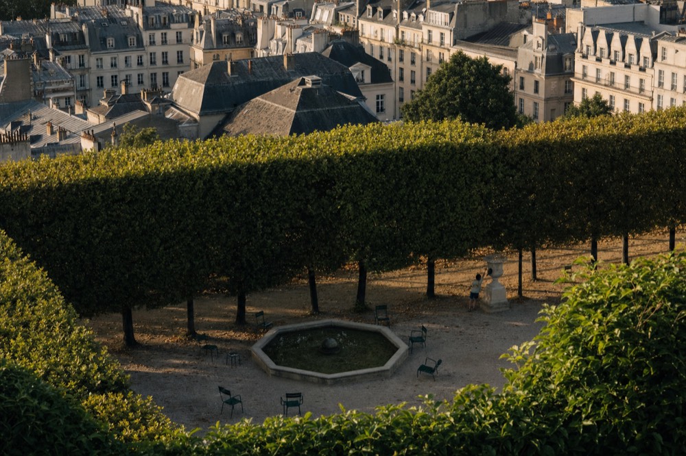 Elevated view of a Paris garden courtyard with clipped trees, an octagonal basin, empty green chairs and mansard rooftops beyond.