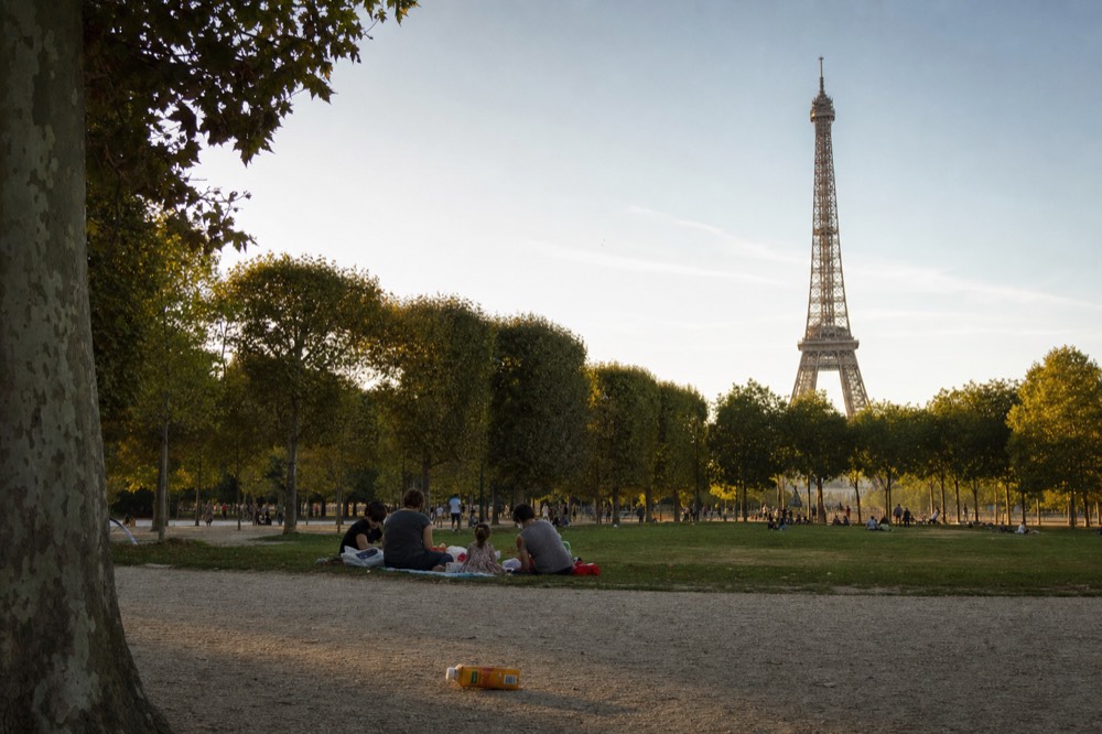 A family sits on a park lawn at Champ de Mars with plane trees and the Eiffel Tower rising in the distance.