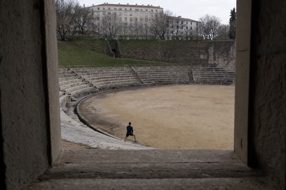 Se ven gradas de anfiteatro de piedra y una arena de arena a través de una apertura oscura, con un visitante lejano y bloques de apartamentos al fondo.
