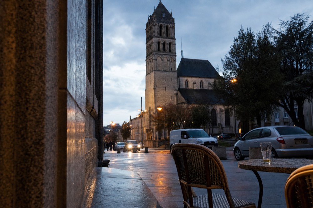 Se alza la torre campanario románica de una iglesia tras el pavimento mojado, coches aparcados, árboles y sillas de café con un vaso sobre una mesa.