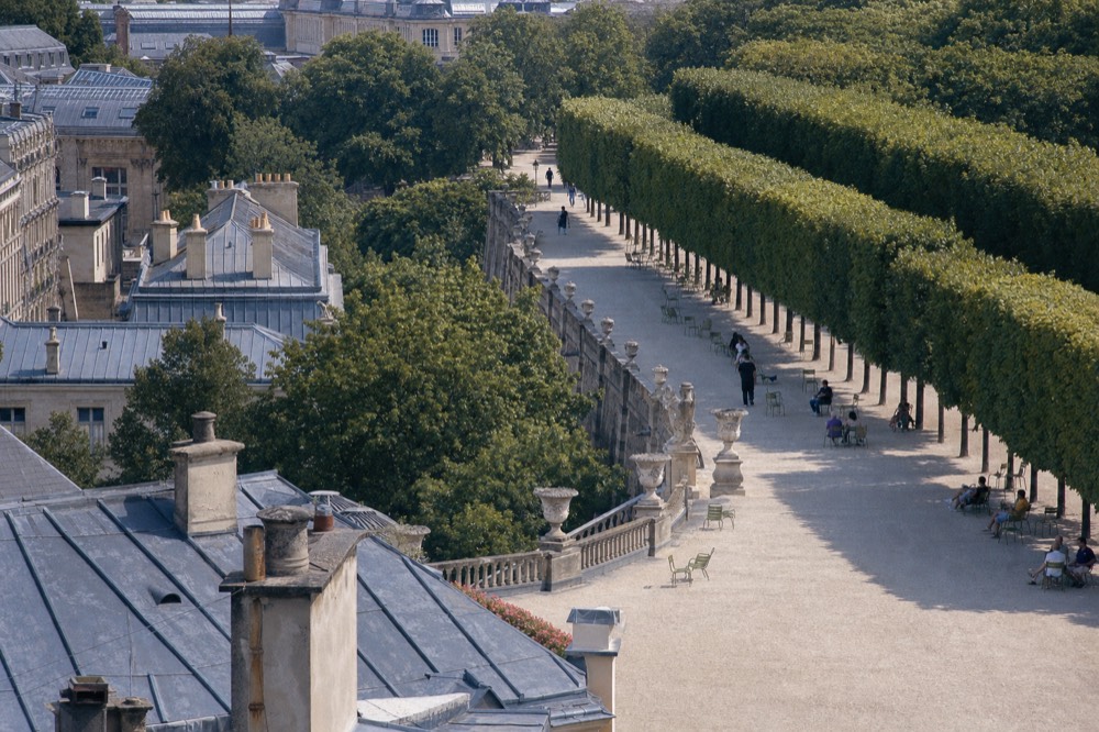 Vista elevada sobre los tejados de París hacia un callejón de jardín de grava bordeado de árboles podados, sillas verdes, urnas y paseantes a lo lejos.