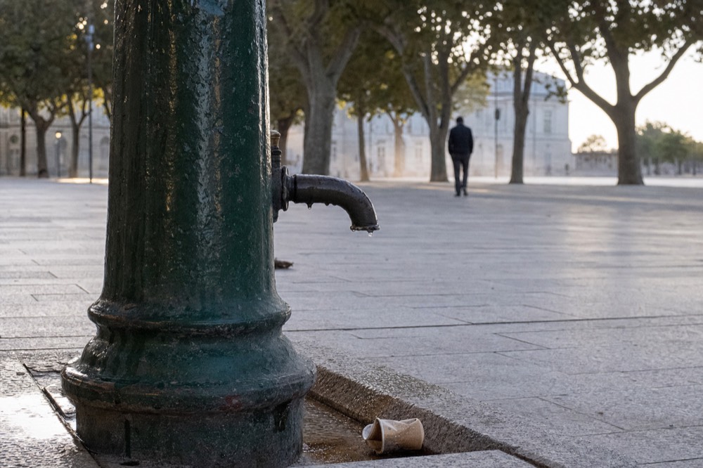 Primer plano de un caño de fuente de agua potable verde oscuro goteando sobre un empedrado de piedra, con árboles y un transeúnte al fondo.