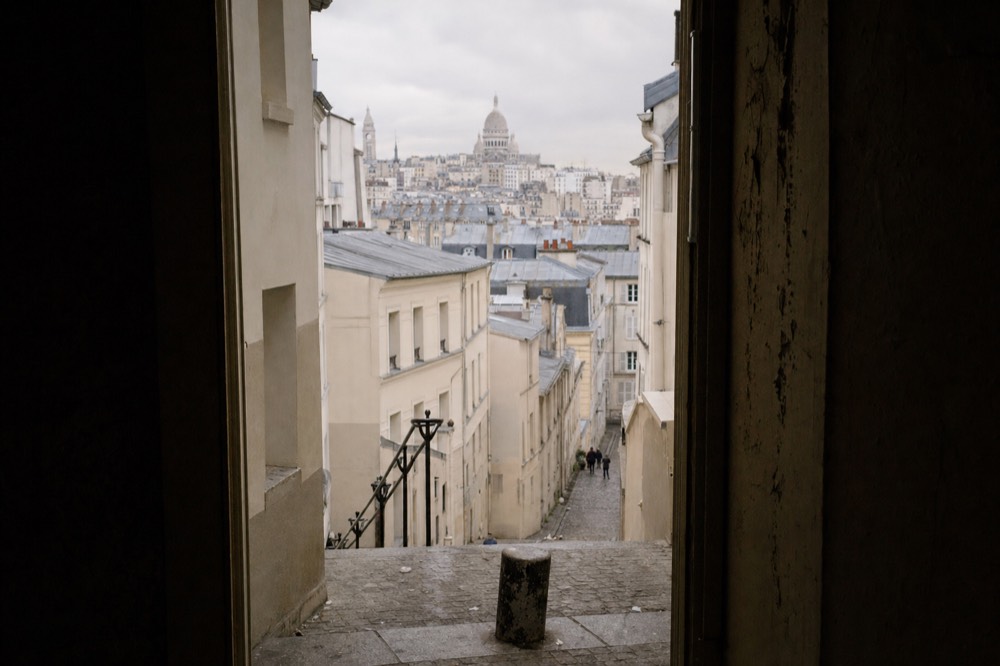 Escaleras de piedra empinadas de Montmartre descienden entre edificios de color crema, con tejados grises y la cúpula del Sacré-Cœur bajo un cielo nublado.