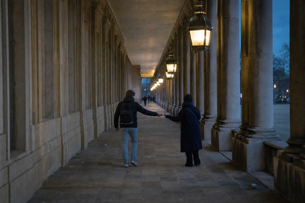 Dos personas intercambian un pequeño objeto bajo una larga arcada de piedra iluminada al atardecer, con columnas, adoquines y caminantes lejanos.