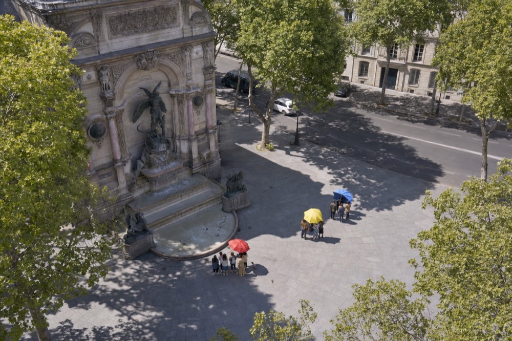 Pequeños grupos se reúnen bajo paraguas rojos, amarillos y azules en una plaza empedrada de París, junto a una gran fuente de piedra y plátanos de sombra.