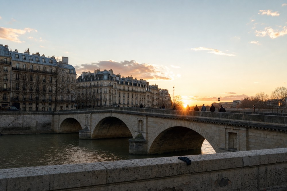 Pequeñas figuras cruzan un puente de piedra de París sobre el Sena al atardecer, con bloques de apartamentos claros y un guante olvidado en el pretil.