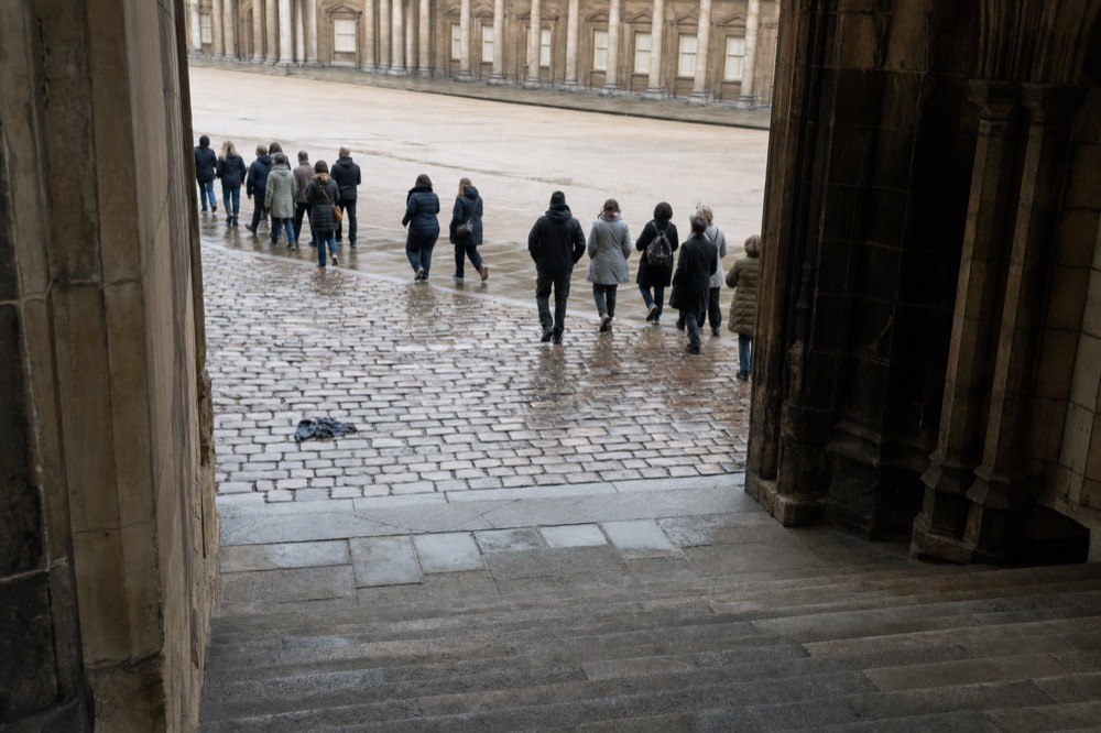 Visitors walk down stone steps through a Gothic arch into a wet cobbled Louvre courtyard, with a dropped scarf on the ground.