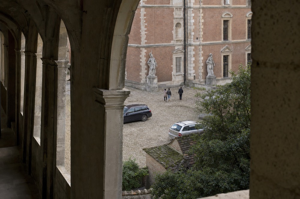 Arcaded stone walkway overlooking a cobbled museum courtyard with statues, brick walls, two cars, and two distant people.