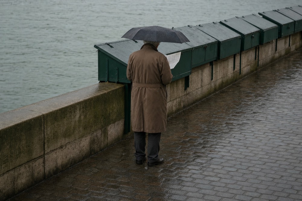 A person with an umbrella reads a paper beside green riverside boxes on a wet cobbled quay.