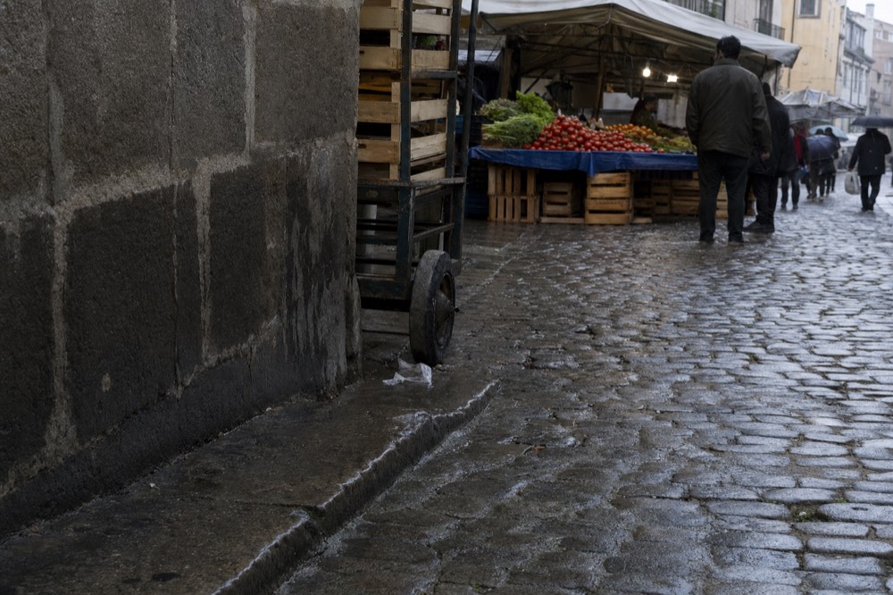 Wet cobblestone Paris market street with a produce stall under an awning, crates of tomatoes, a handcart wheel, and shoppers walking away.