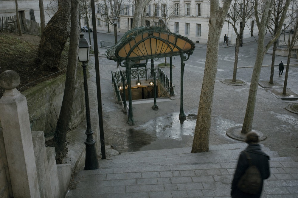 Green Art Nouveau Paris metro entrance with amber canopy at the foot of wet stone steps, bare trees, and a blurred commuter.