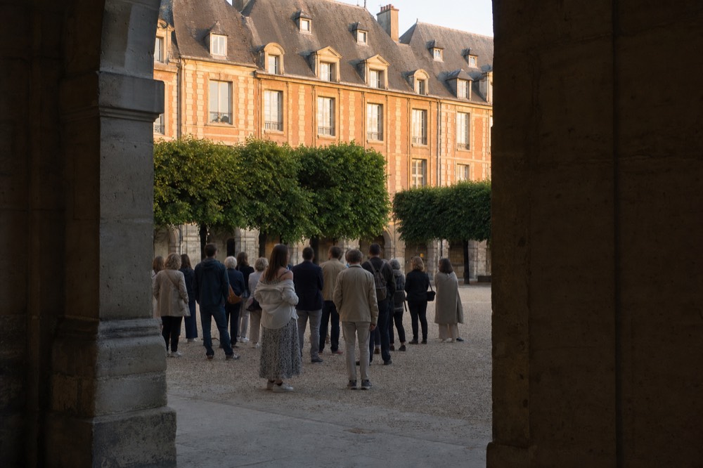 A walking group stands on a gravel Paris square seen from a shaded stone arcade, with clipped trees and brick facades behind.