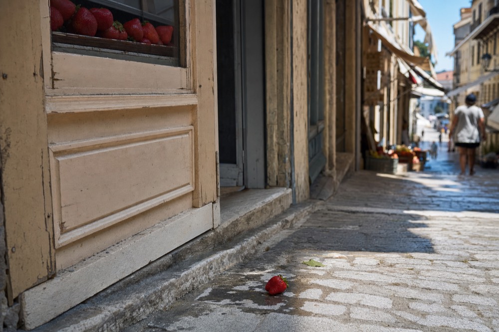 A dropped strawberry lies on damp cobbles beside a worn shopfront on a narrow sloping market street with awnings and distant pedestrians.